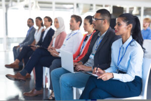 A diverse group of people sat in a row looking ahead, listening to a presentation and taking notes